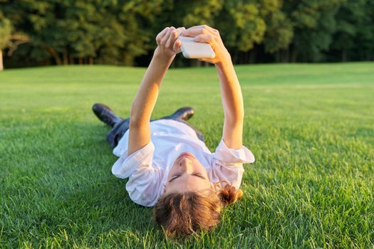 Guy teenager lying on grass with smartphone. Male in white T-shirt with phone in his hands, top view, green lawn background