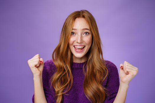 Optimistic, lucky young attractive female student winning trip around european smiling broadly from success and delight clench fists in triumph and celebration gesture, happy to win over purple wall
