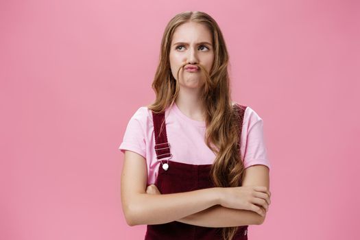 Portrait of cute funny and charismatic girl making moustache from hair and silly face fooling around mimicking seriousness holding hands crossed against chest and looking left strict and serious