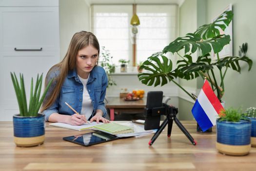 Young female university student sitting at home studying online, looking at smartphone webcam. On table flag of Netherlands, education in Holland, e-education, e-learning, technology, knowledge