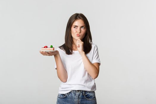 Thoughtful smiling pretty girl pondering while holding piece of cake, white background