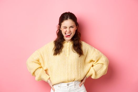 Sassy beautiful young woman, looking determined and self-assured, showing tongue, striking pose, standing against pink background