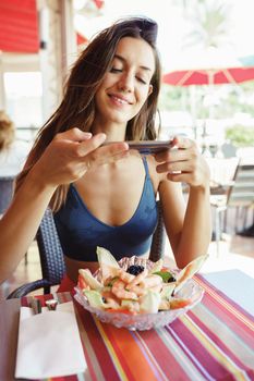 Young woman photographing her salad with a smartphone while sitting in a restaurant