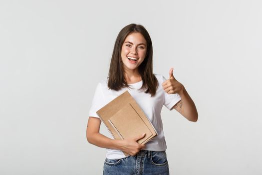 Cheerful smiling girl holding notebooks and showing thumbs-up, recommend courses or school