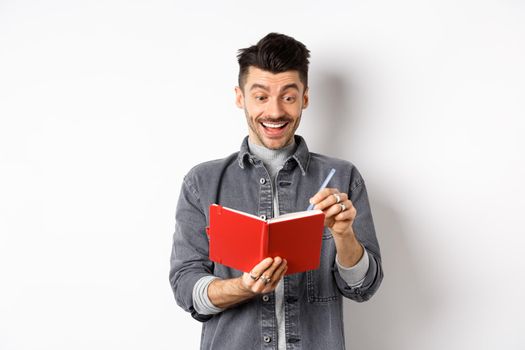 Excited guy writing in planner and smiling, writing down ideas in journal or diary, standing on white background
