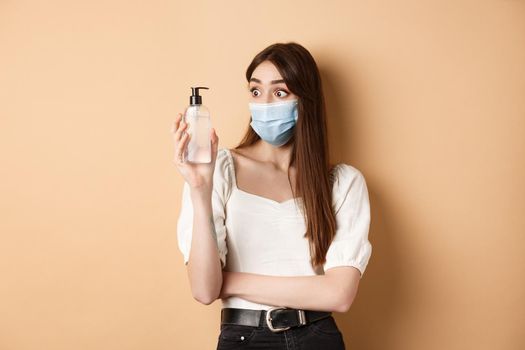 Covid-19 and preventive measures concept. Excited girl in medical mask look amazed at hand sanitizer bottle, standing on beige background