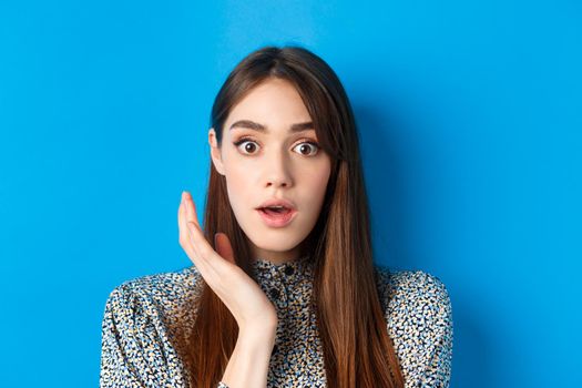 Candid girl with long hair and makeup, looking shocked and surprised, open mouth and stare speechless at camera, standing on blue background