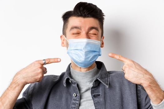 Covid-19, health and lifestyle concept. Cheerful smiling man pointing at sterile medical mask, protect himself from corona outbreak, standing on white background