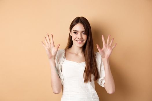Attractive young woman show fingers number ten, smiling and looking confident, standing on beige background