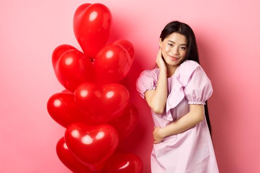 Beautiful asian girl in dress smiling coquettish, flirting on valentines day, looking sensual at camera, posing near valentines heart balloons, pink background