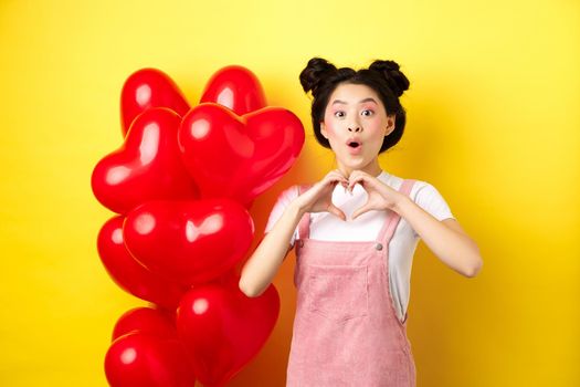 Surprised asian woman looking in awe, showing heart gesture, making love confession on valentines day, standing near romantic red balloons over yellow background