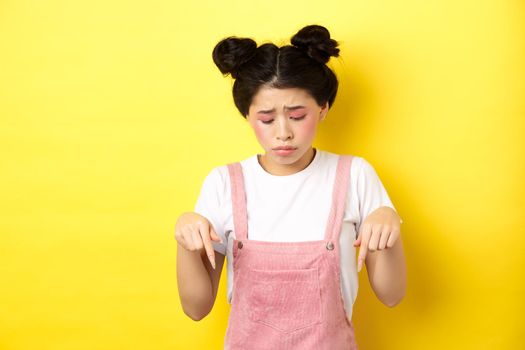 Sad miserable girl with glamour bright makeup, looking and pointing down upset, showing bad thing, standing against yellow background
