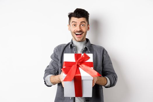 Surprised young man smiling excited, holding big gift box on valentines day holiday, receive surprise present, standing amazed on white background