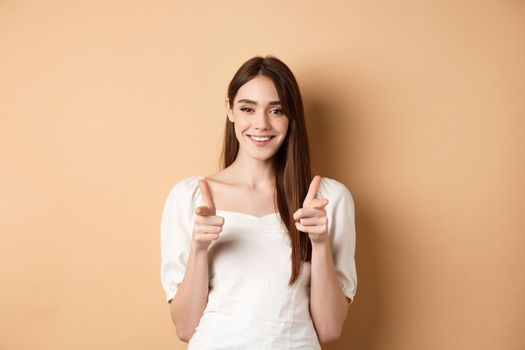 We need you. Smiling young woman pointing fingers at camera to beckon or invite people, standing on beige background