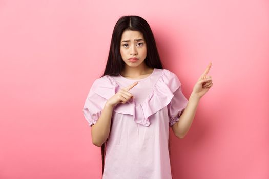 Sad and distressed asian girl frowning, sulking and pointing fingers right at disappointing bad news, feeling unhappy, standing against pink background
