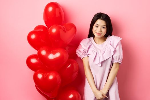 Cute asian girl in dress looking shy and smiling, standing modest near valentines day balloons, blushing on romantic date, looking at camera, pink background