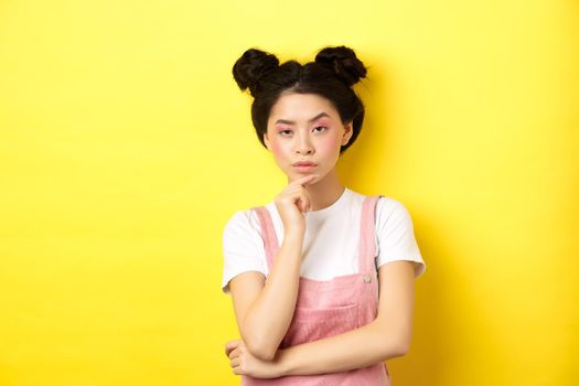 Suspicious asian girl looking at camera and thinking, stare with disbelief, standing with makeup on yellow background