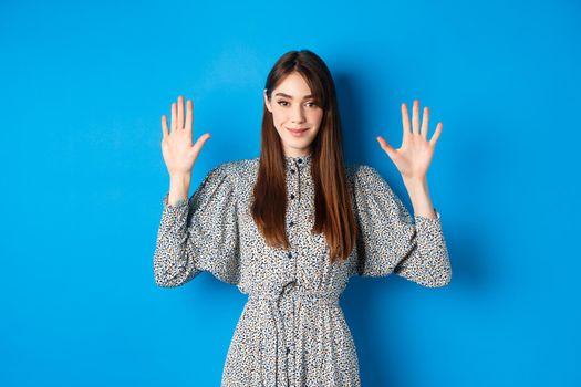 Young beautiful girl in dress with natural long hair, showing number ten with fingers and smiling, standing against blue background