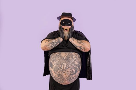 Shocked plump man in hero costume holds cheeks standing on purple background