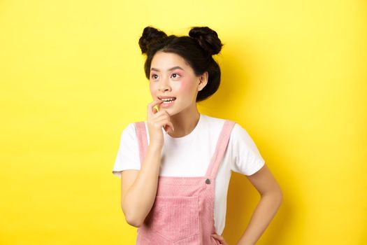 Pensive asian girl feeling excited, biting fingernail and looking left thoughtful, making choice, standing in summer clothes against pink background