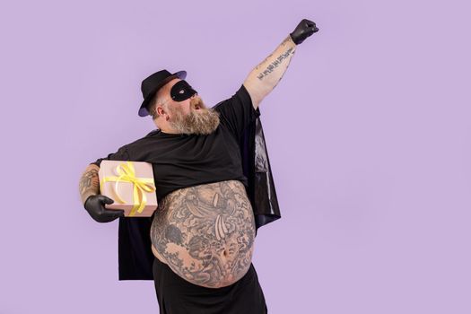 Funny gentleman with overweight wearing hero costume holds present on purple background