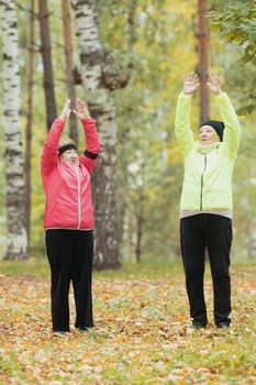 Mature woman in colourful jackets doing gymnastics in an autumn park after a scandinavian walk. Arms up