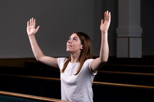 A Christian girl in white shirt is sits with his hands up and face and praying with humble heart in the church