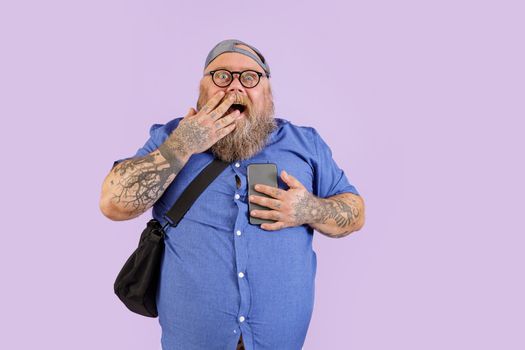 Surprised obese man holds mobile phone covering mouth by palm on purple background