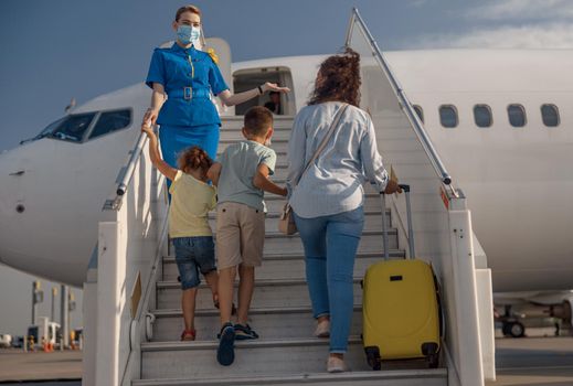Back view of mother with two little kids and suitcase boarding the plane. Air stewardess wearing protective mask welcoming the family