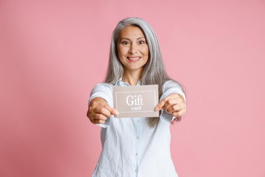 Attractive grey haired Asian lady holds gift card on pink background