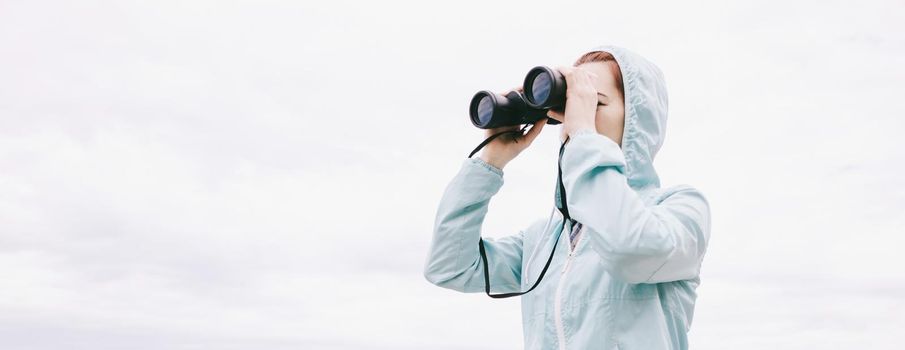 Explorer woman looking through binoculars in front of sky.