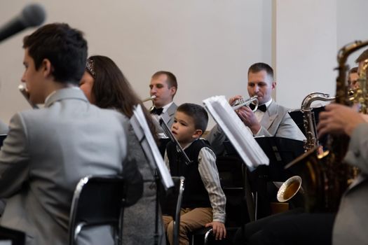 KOROSTEN - NOV, 10, 2019: A little boy sits in an orchestra near the musicians play different musical instruments in gray suits