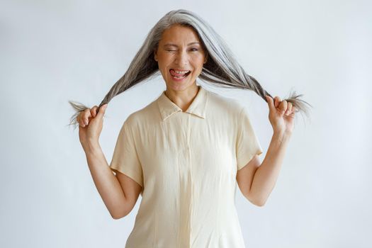 Middle aged Asian lady grimaces and holds long silver hair on light grey background