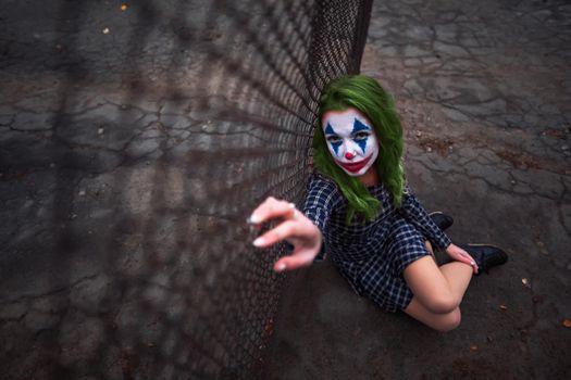 Greenhaired girl in chekered dress with joker makeup sitting near wire mesh fence on the ground.