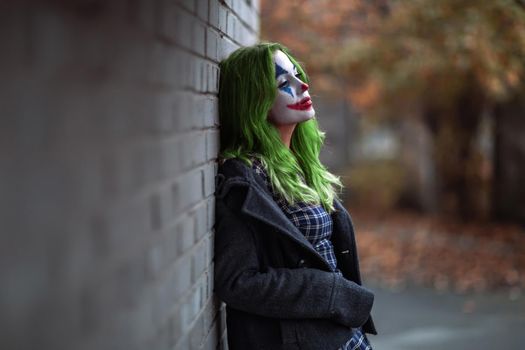 Portrait of a greenhaired girl in chekered dress with joker makeup on a brick wall background.