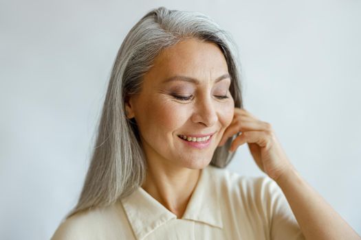 Attractive smiling grey haired lady in stylish shirt touches cheek on light background