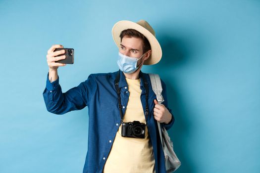 Covid-19, pandemic and travel concept. Male tourist in straw hat and medical mask recording video on smartphone during vacation, taking pics on summer holiday, blue background