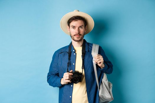 Handsome young tourist in summer hat, backpacking and travelling, holding photo camera, standing on blue background