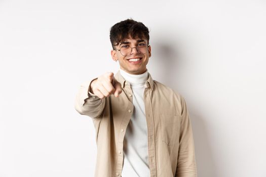 Handsome modern guy in glasses pointing at camera, smiling and choosing you, recruiting or inviting to event, standing on white background