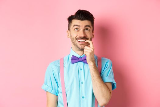 Image of smiling man making choice, looking dreamy and happy up, biting fingernail with tempted expression, wanting something, standing over pink background