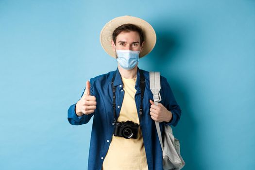 Covid-19 and summer holidays concept. Smiling guy travelling in medical mask and straw hat, backpacking and showing thumbs up, approve and praise travel agency, blue background