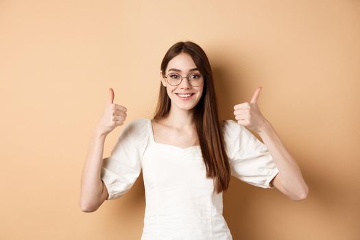 Happy girl in glasses show thumbs up, recommend eyewear store, smiling satisfied, standing on beige background
