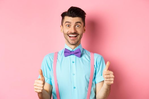 Excited caucasian guy showing thumbs up and praising great work, well done gesture, standing in suspenders and bow-tie on pink background.
