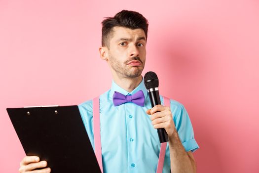 Holidays concept. Funny man with bow-tie making speech, perform on party events, holding script on clipboard and microphone, entertaining you, standing over pink background