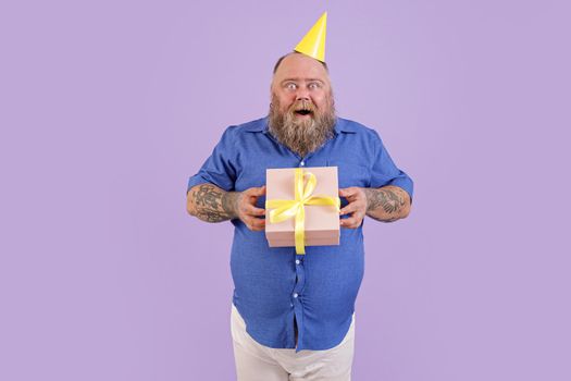 Excited plump man wearing tight shirt holds present on purple background