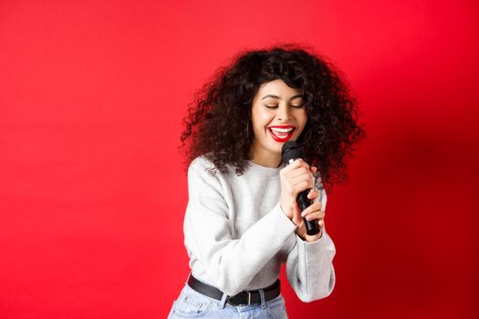 Hobbies and leisure concept. Beautiful woman singing in microphone and smiling, having fun at karaoke, standing on red background.
