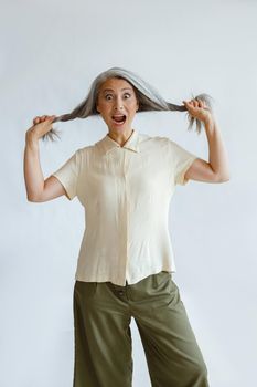 Shocked Asian woman holds hoary hair locks on light grey background