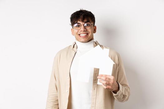 Real estate and mortgage concept. Cheerful young guy renting apartment, showing paper house cutout and smiling happy, buying property, white background