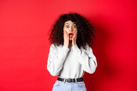 Portrait of shocked woman scream amazed, touching face and looking at camera at impressive promo offer, standing in sweatshirt on red background