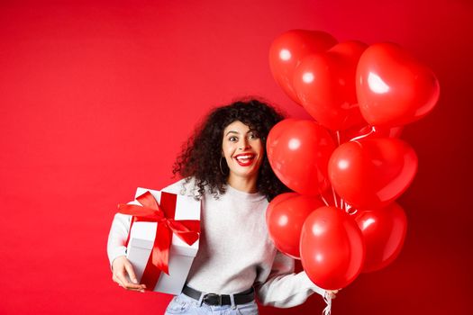 Holidays and celebration. Happy birthday girl holding gift and posing near party helium balloons, smiling excited at camera, red background